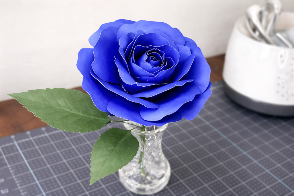 Blue paper rose in a glass vase with green leaves